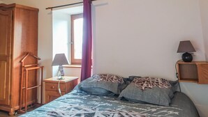 A bedroom with a bed, a wooden wardrobe, a nightstand with a lamp, a window with curtains, and a mirror.