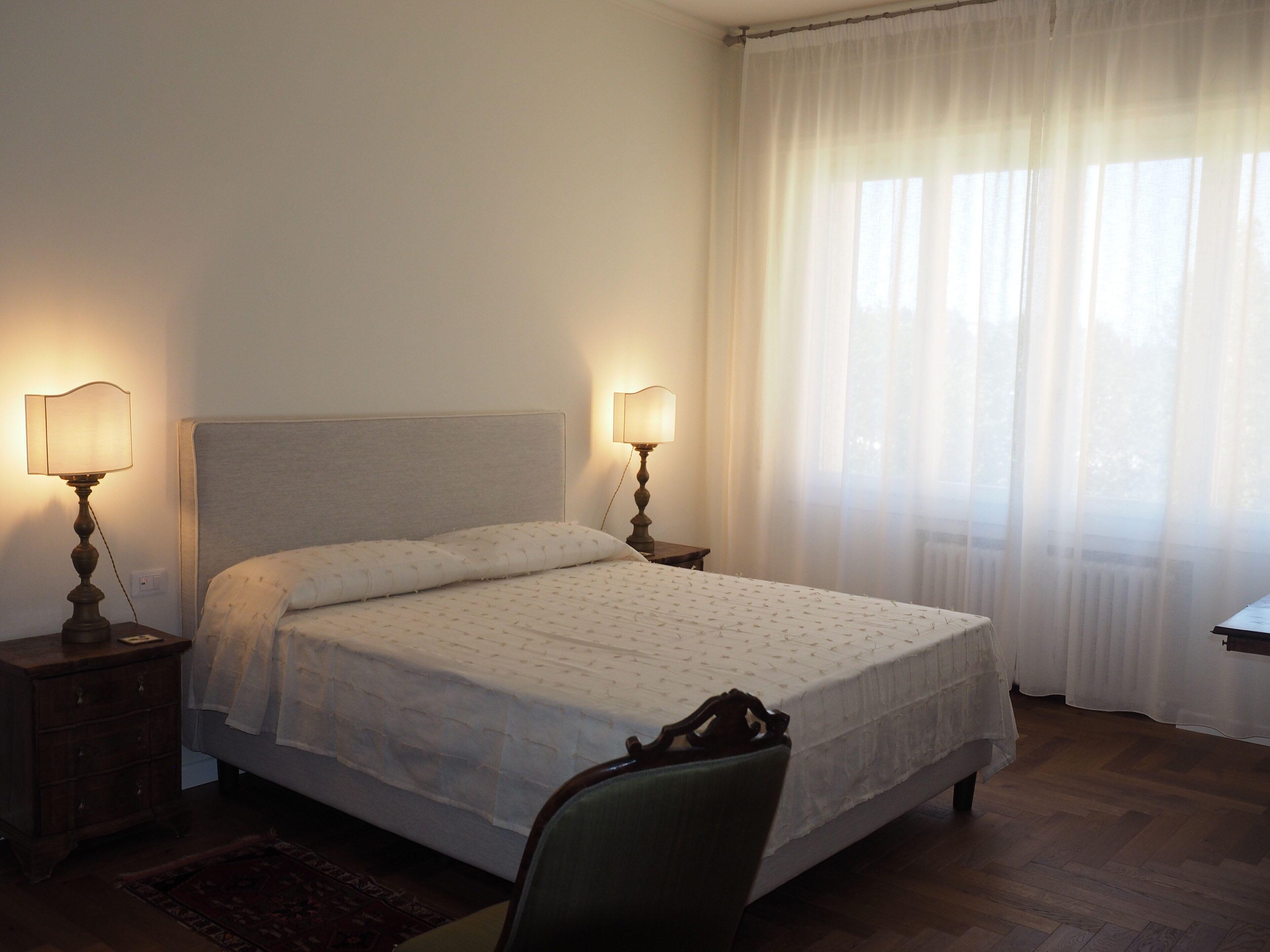 A bedroom with a bed, two bedside lamps, a chair, a wooden dresser, and a window with sheer curtains.