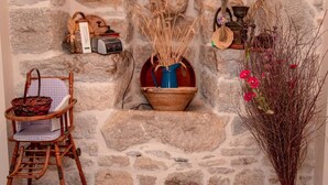 Una pared de piedra rústica con una silla de madera, un florero azul con plantas secas y un jarrón con flores rojas.