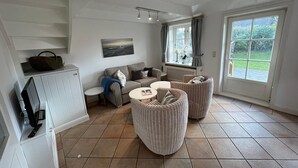 Un vaste espace de vie comprenant un canapé, deux fauteuils, une table basse et un radiateur. On y trouve une fenêtre avec des rideaux, un tableau accroché au mur et un panier posé sur une étagère.