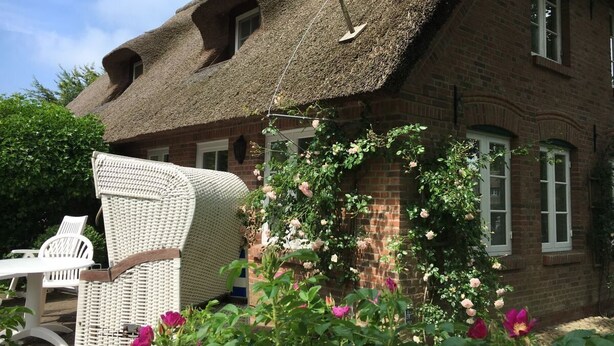 Une maison traditionnelle en briques, avec un toit de chaume, une chaise en osier blanche et des fleurs en pleine floraison dans le jardin.