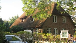 Une maison traditionnelle en briques, avec un toit de chaume, entourée de verdure et d’un mur de pierres.