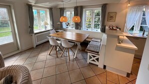 Un coin repas avec une table en bois, des chaises blanches et un banc. On y trouve deux lampes suspendues, un radiateur et une fenêtre avec des rideaux.