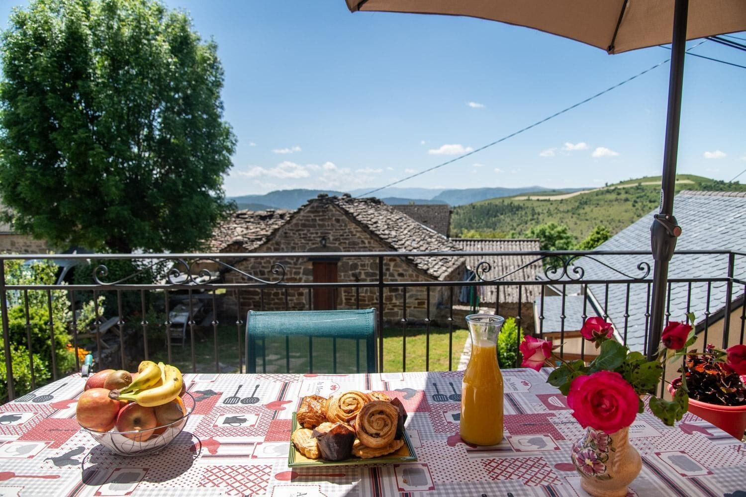 A table with food and drinks, a potted plant, and a view of a stone building and hills.