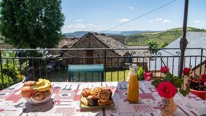 A table with food and drinks, a potted plant, and a view of a stone building and hills.