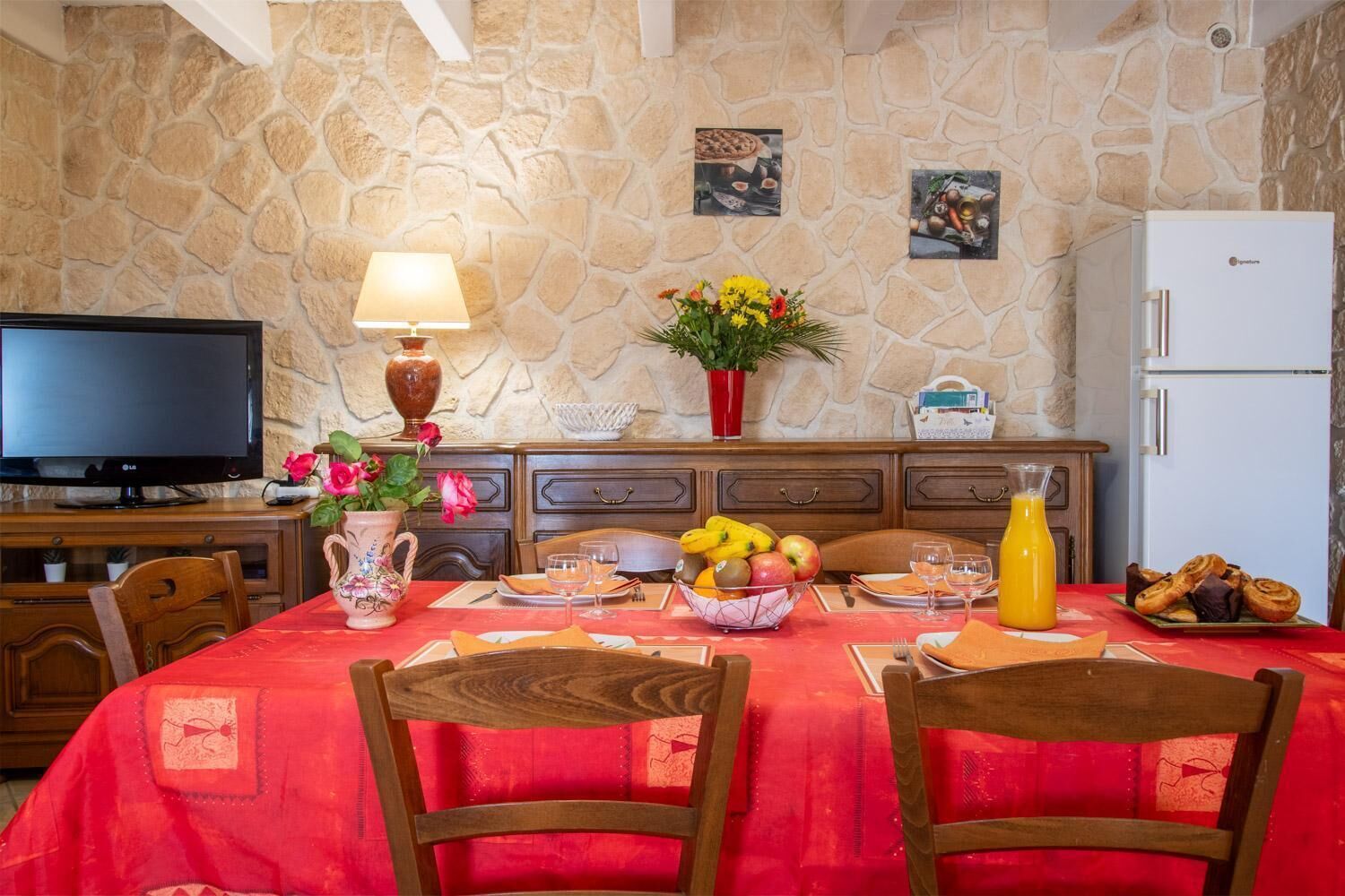 Un coin repas avec un mur en pierre, un téléviseur, une lampe, un vase de fleurs, un panier de fruits et un réfrigérateur.