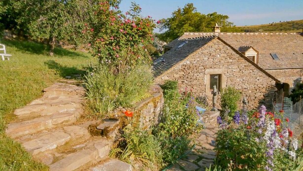 Una cabaña de piedra con techo de tejas, rodeada de un jardín con diversas plantas y flores.