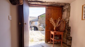 Una entrada con suelo de piedra y una puerta de madera que da a un patio, una cesta de mimbre sobre una mesita y un muro de piedra.