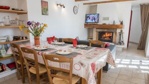 Un coin repas avec une table dressée pour quatre personnes, entourée de chaises en bois. On aperçoit une cheminée au fond, et un téléviseur est fixé au-dessus.