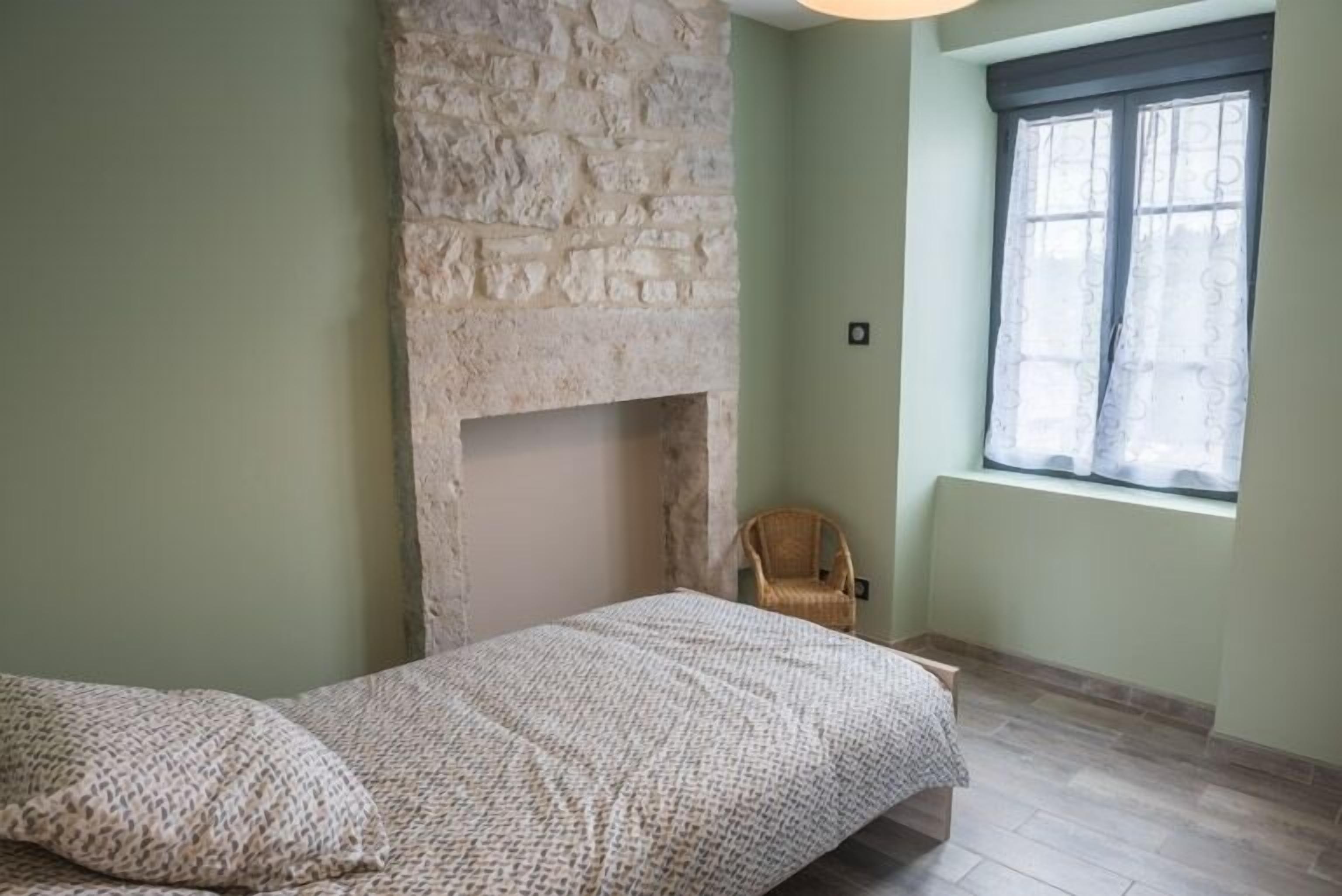 Une chambre avec une cheminée en pierre, un lit recouvert d’une couvre-lit à motifs, un fauteuil en osier et une fenêtre avec des rideaux.
