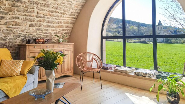 A cozy room with a stone wall, a large arched window, a wooden cabinet, a wicker chair, and a sofa with cushions.
