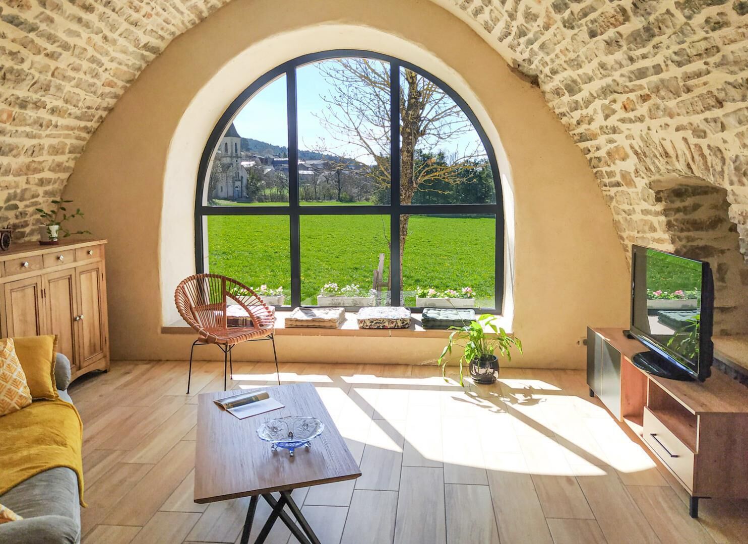 Ein gemütliches Zimmer mit einem großen Bogenfenster, einem Holztisch, einem Korbstuhl und einem Fernseher.