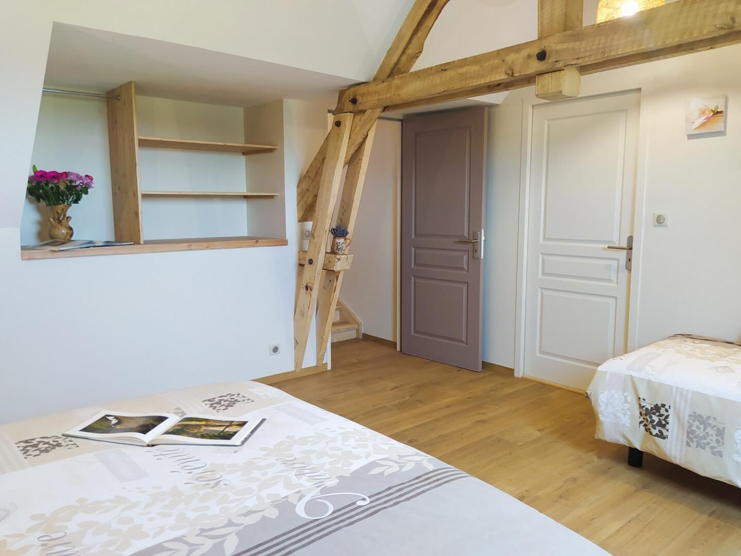 A bedroom with a wooden ceiling, a bed with a patterned bedspread, a small shelf with a vase of flowers, and a door leading to another room.