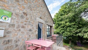 Un bâtiment en pierre avec une enseigne indiquant « Gîte Lozère », un mur en pierre, une porte bleue, une table et des chaises roses, et un arbre en arrière-plan.