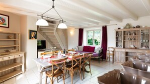 Un coin repas avec une table blanche, des chaises en bois et un lustre. On aperçoit un escalier au fond, qui mène à un autre niveau.