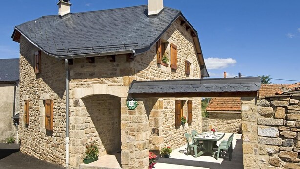 Un bâtiment en pierre avec un toit en pente, un patio couvert avec une table et des chaises, et une petite arche.
