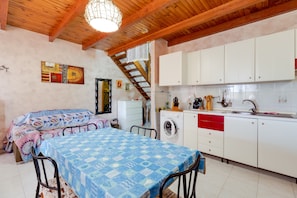 A bedroom with a bed, a table, and a washing machine in a room with a kitchen area.