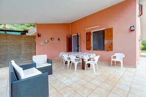 Eine überdachte Terrasse mit Tisch, Stühlen und einer Rattan-Couch, sowie eine Tür, die in einen Außenbereich führt.