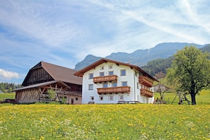 Un bâtiment à deux étages, doté d’un toit en pente et de balcons en bois, entouré d’un champ de fleurs jaunes et avec des montagnes en arrière-plan.