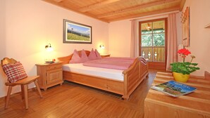 Een houten bed met een geruite plaid, een nachtkastje met een lamp, een stoel met een ruitkussen, een ingelijst schilderij aan de muur, en een raam met gordijnen.