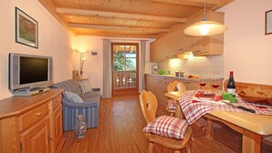 Een gezellige kamer met een televisie, een bank, een eettafel, stoelen en een keukenhoek met kastjes en een raam.