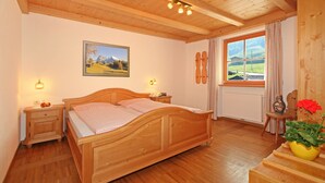 Een slaapkamer met houten frame, met een bed, nachtlampen, een raam met gordijnen, een schilderij aan de muur en een vaas met bloemen op een tafel.