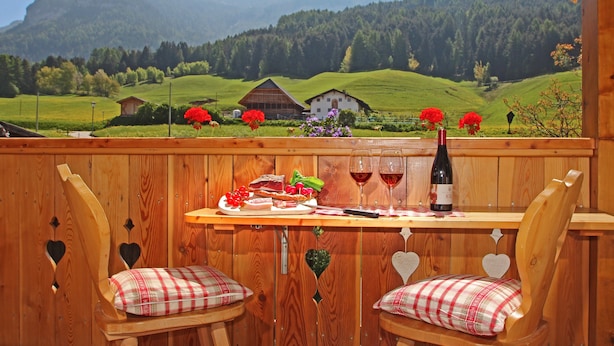 Een houten terras met een tafel gedekt voor twee personen, inclusief een fles wijn, glazen, een bord met eten en een prachtig uitzicht op bergen en huizen.