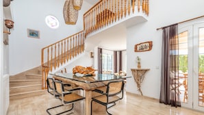 Un comedor espacioso con una mesa y sillas de madera, una escalera con barandillas de madera y vistas a través de un ventanal.