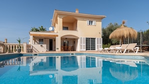 Una casa de dos pisos con piscina, un patio con sillas de descanso y una sombrilla de paja.
