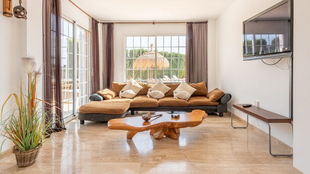 Una sala de estar moderna con un sofá seccional, una mesa de centro de madera, un televisor montado en la pared y ventanas amplias con cortinas.
