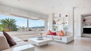 A modern living room with a white sofa, a coffee table, and a TV. There is a large window with a view of the city and mountains. - Villa 'Berganti' with Private Pool, Wi-Fi, and Air Conditioning (Canet de Mar)