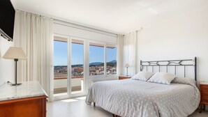 A bedroom with a bed, a nightstand, a lamp, and a view of a cityscape through a large window. - Villa 'Berganti' with Private Pool, Wi-Fi, and Air Conditioning (Canet de Mar)