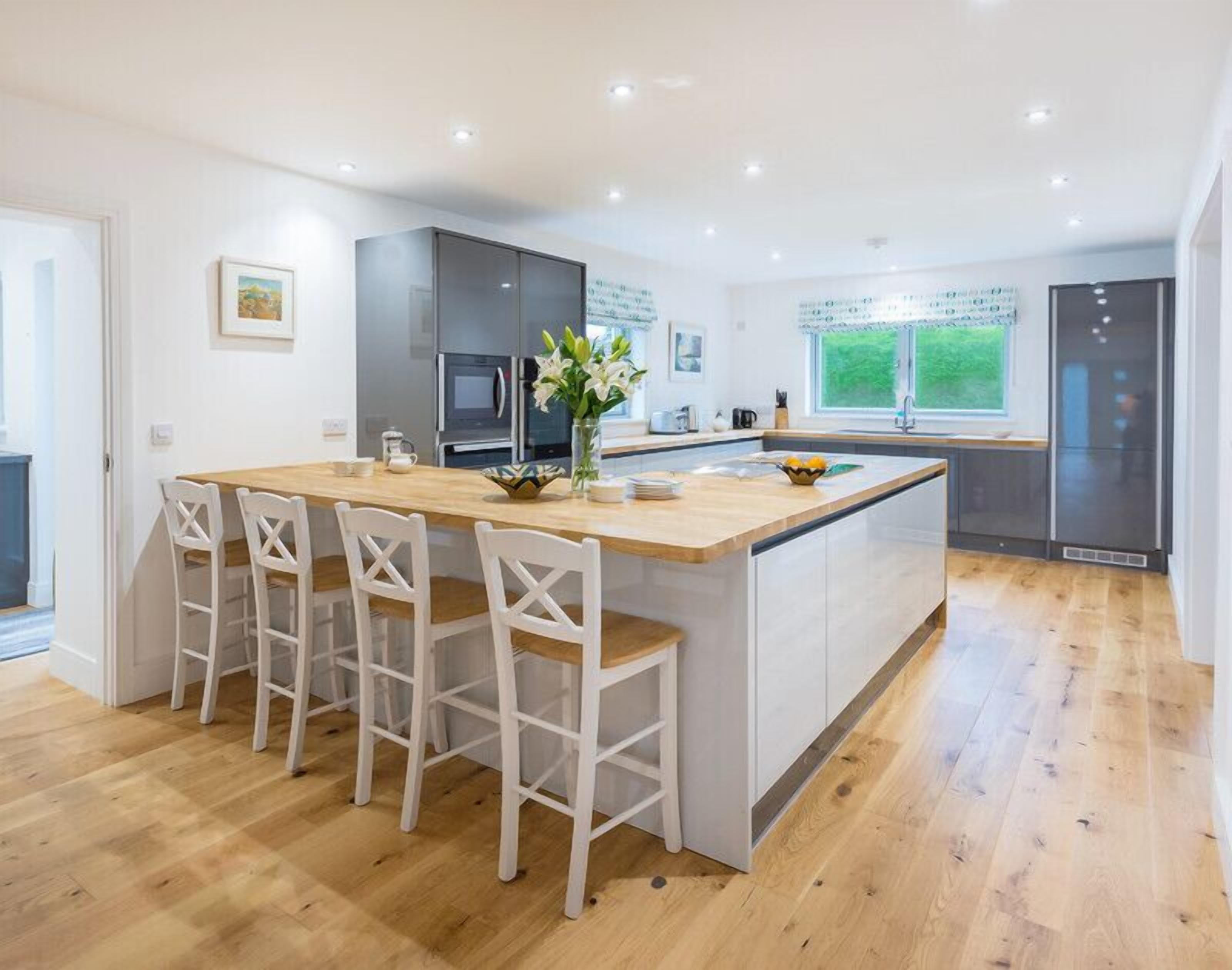 A modern kitchen with a central island, white bar stools, a refrigerator, and a window with a view of greenery.