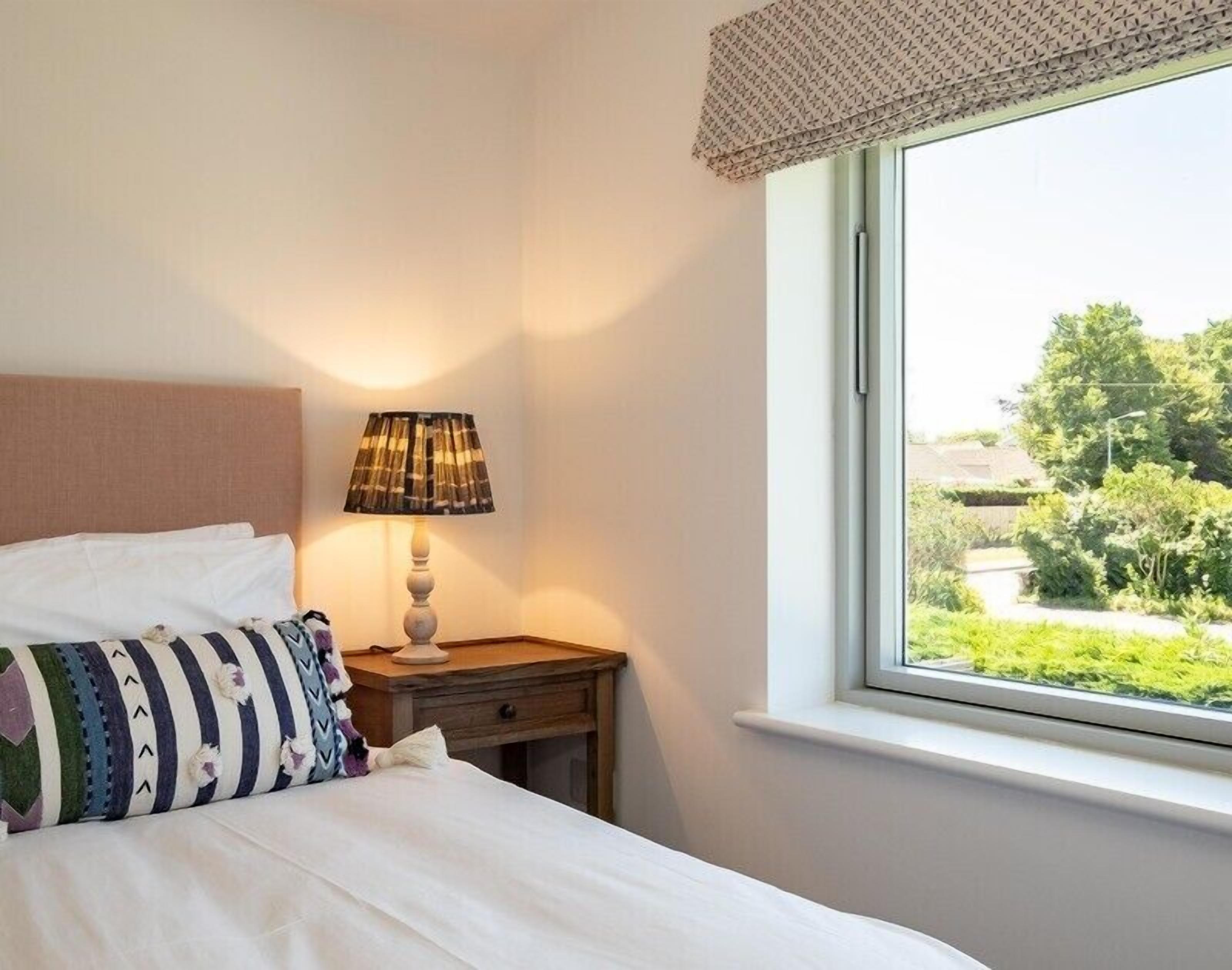 Un lit bien fait, agrémenté d’un coussin à motifs, d’une lampe de chevet et d’une fenêtre donnant sur un paysage verdoyant.