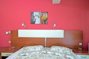 A bedroom with a wooden headboard, a bed with floral bedding, a red wall, and a painting of a canal scene.