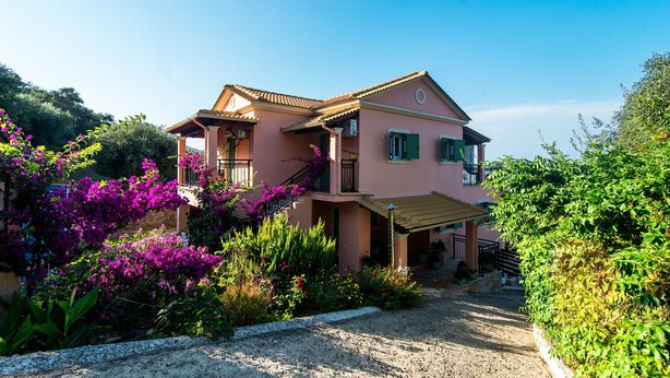 Una casa de dos pisos con un balcón, rodeada de abundante vegetación y plantas con flores.
