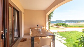 An outdoor dining area with a wooden table and chairs, a view of the sea and hills, and a door leading to a covered area.
