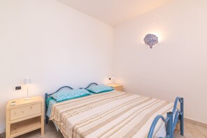 Une chambre à coucher comprenant un lit, deux tables de chevet, une petite table, un luminaire fixé au mur et une chaise bleue.
