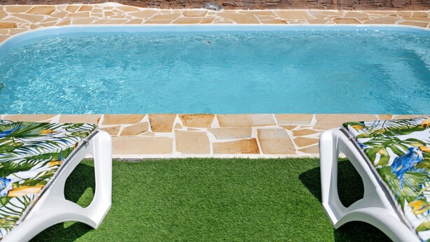 Une piscine aux bords recouverts de pierres, une pelouse et deux chaises longues blanches ornées d’un motif tropical.