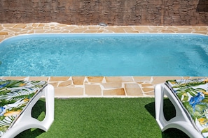 A swimming pool with stone tiles, a grassy area, and two white lounge chairs with a tropical print.