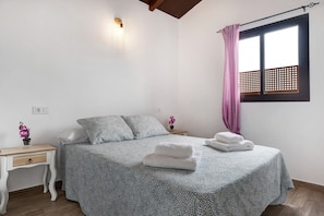 A bedroom with a bed, a nightstand, a wall-mounted light, a window with a curtain, and a small table with a flower vase.