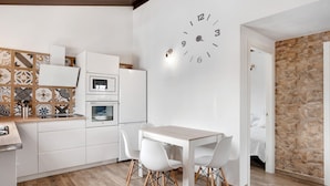 Une cuisine moderne avec des meubles blancs, un four encastré, une table à manger avec des chaises blanches et une grande horloge murale.