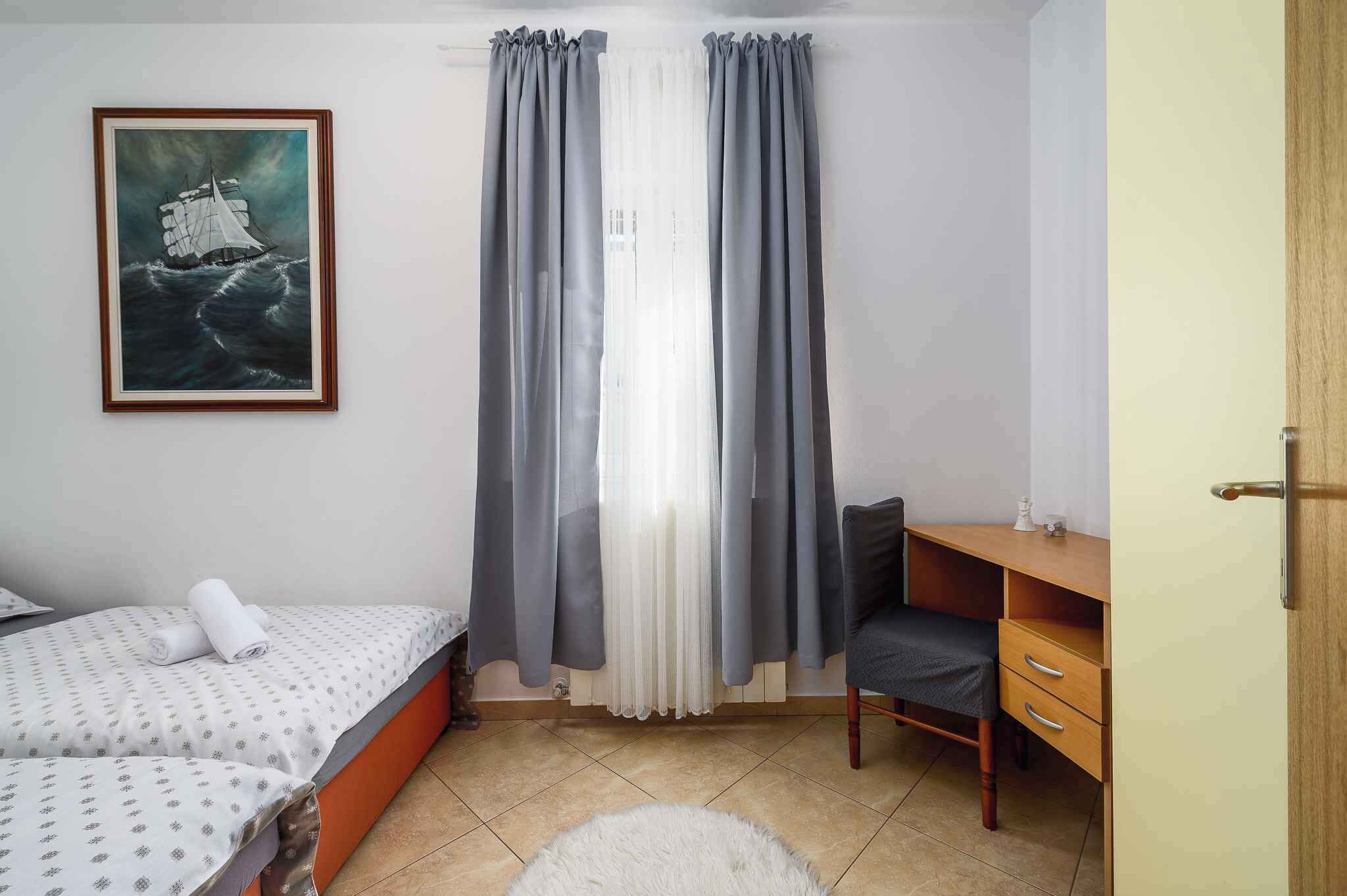 Un dormitorio con cama, escritorio y silla. Hay un cuadro en la pared y una ventana con cortinas.