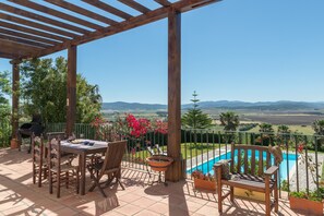 Un patio cubierto con mesa y sillas, una parrilla de barbacoa, una piscina y vistas al campo.