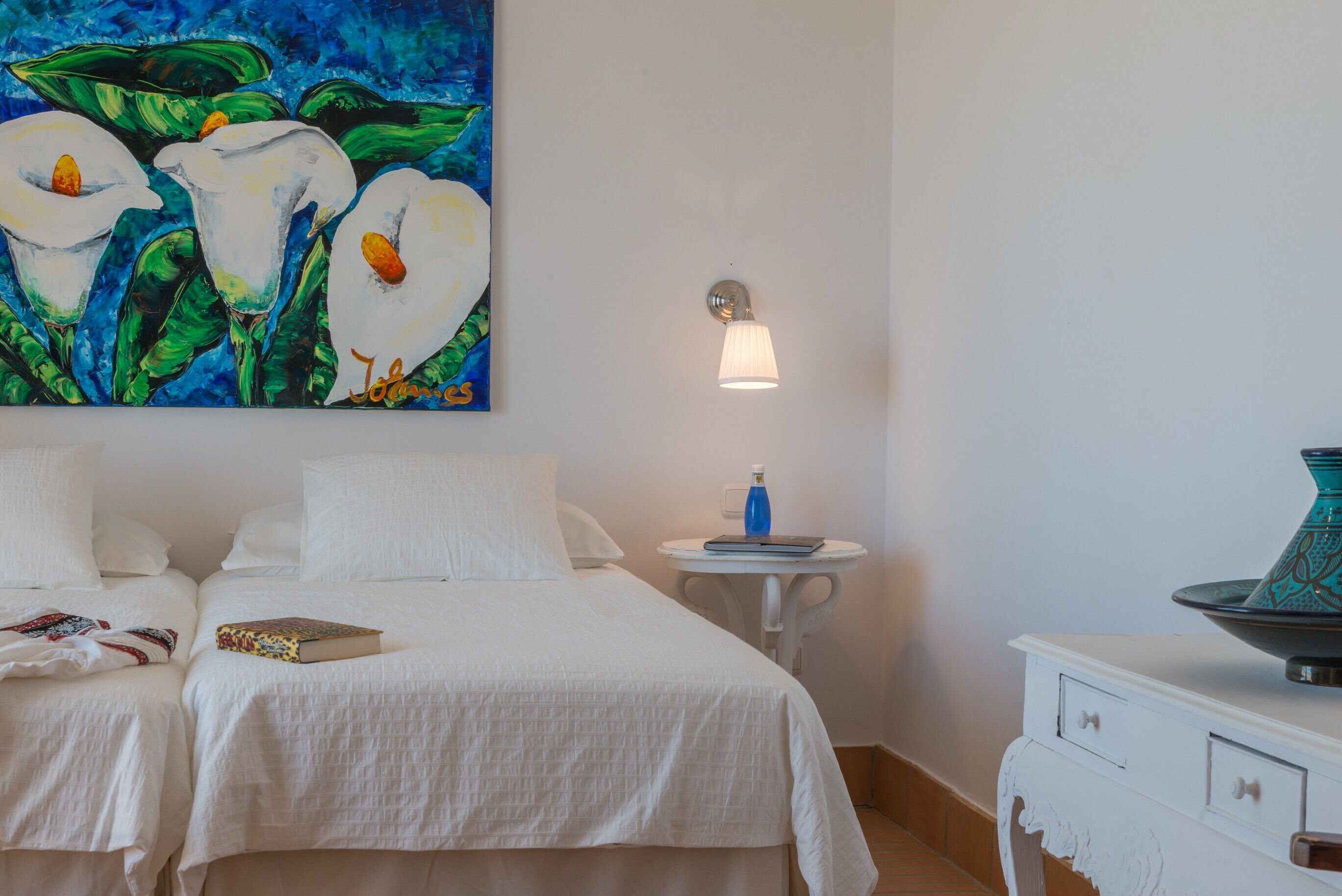 Una habitación de hotel con dos camas, un cuadro de flores, una lámpara de pared, una mesita con una botella y un jarrón decorativo.