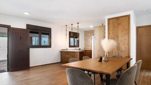A modern dining area with a wooden table, grey upholstered chairs, a vase with feathers, and a kitchen in the background. - Holiday Home 'Casa La Teja 2' with Pool and Wi-Fi (Vejer de la frontera)