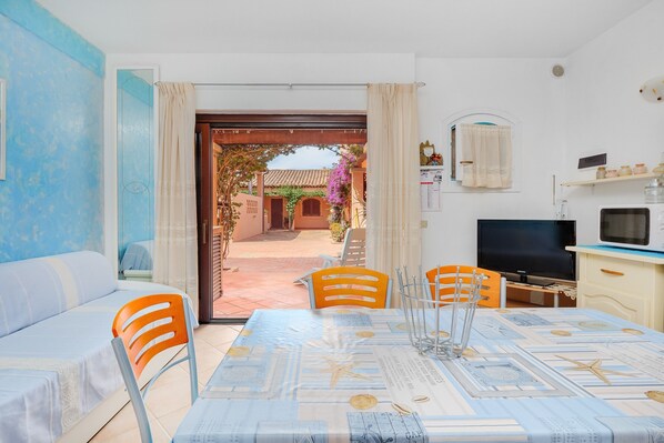 A room with a dining table, chairs, a sofa, a TV, and a view of an outdoor area with a gate and plants.