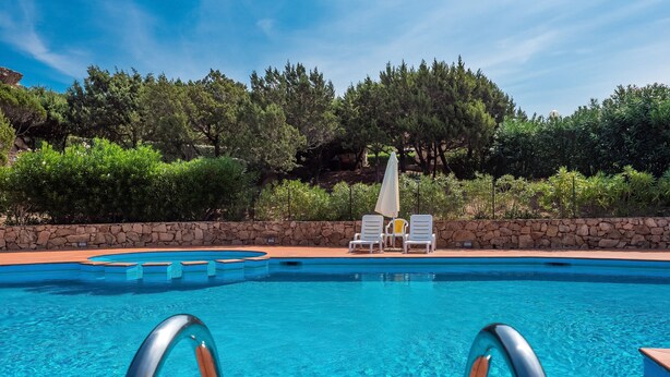A swimming pool with clear water, surrounded by a stone wall, with lounge chairs and umbrellas. - Casa Turisarda Apartment with Shared Pool, Terrace, and Air Conditioning (Baja Sardinia)