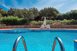 A swimming pool with clear water, surrounded by a stone wall, with lounge chairs and umbrellas.
