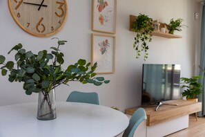 Ruokailutila, jossa on pyöreä pöytä, tuoleja, vihreälehtinen maljakko, seinäkello, kehystettyjä kukkakuvia, televisio ja puinen hylly, jossa on kasveja.